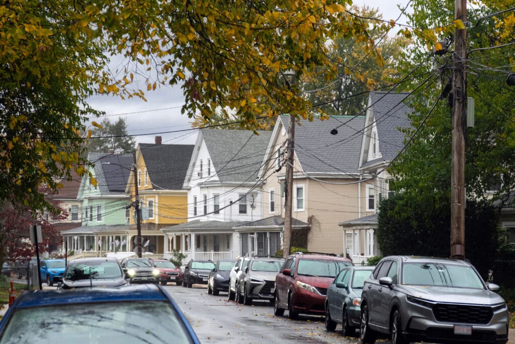 A residential street lined with various two-story houses in shades of yellow, green, and beige. The foreground features parked cars along the road, and power lines run along the street. Trees with yellowing leaves frame the scene.