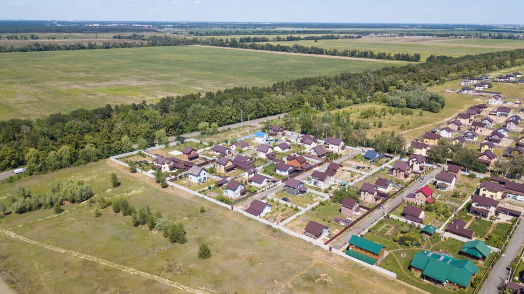 An aerial view of a residential neighborhood featuring several houses with brown roofs. The area includes green spaces and trees, with a large open field in the background, surrounded by flat, agricultural land.