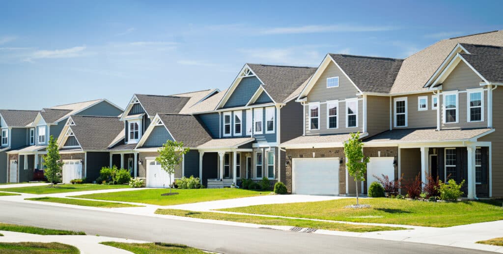 A row of modern single-family homes with various architectural styles, featuring front lawns and driveways. Bright blue sky overhead and green grass in the foreground indicate a suburban neighborhood.